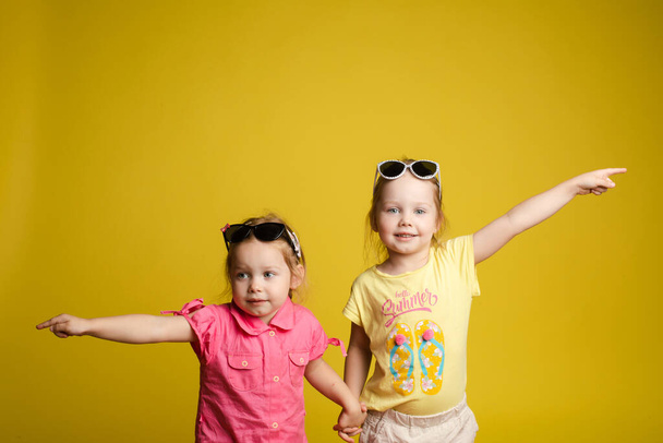 Dwa szczęśliwe piękne stylowe dziewczynki w okularach przeciwsłonecznych pozowanie odizolowane żółte tło studio - Zdjęcie, obraz