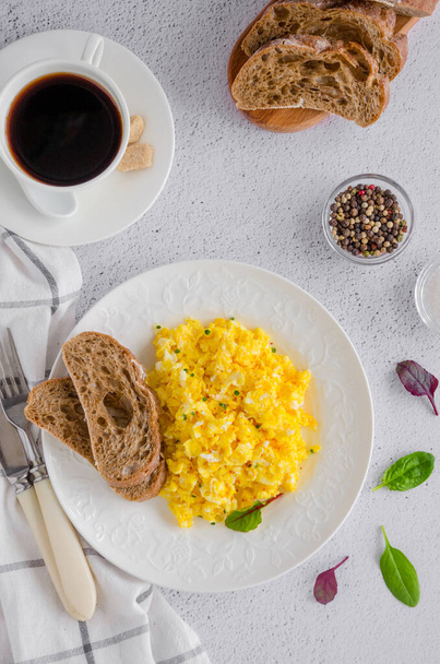 Rührei aus frischen Bio-Eiern auf einem weißen Teller mit Roggenbrot auf hellem Hintergrund und einer Tasse Kaffee. gesundes Frühstück. vertikale Orientierung. Ansicht von oben, Nahaufnahme - Foto, Bild