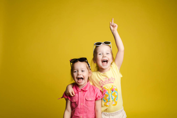 Two happy beautiful stylish little girl wearing sunglasses posing isolated yellow studio background - Foto, Imagen