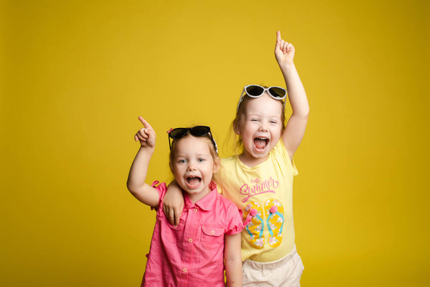Two happy beautiful stylish little girl wearing sunglasses posing isolated yellow studio background - 写真・画像