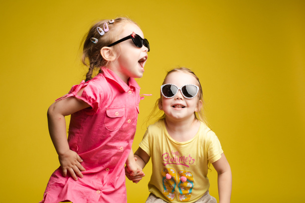 Two happy beautiful stylish little girl wearing sunglasses posing isolated yellow studio background - 写真・画像