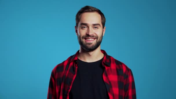 Handsome european man with trendy beard in red plaid shirt on blue studio background. Cheerful guy smiling and looking to camera. - Video