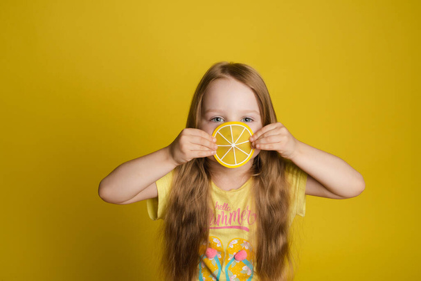 Portrait of smiling little girl holding slice of lemon near eye looking at camera medium close-up - Photo, Image