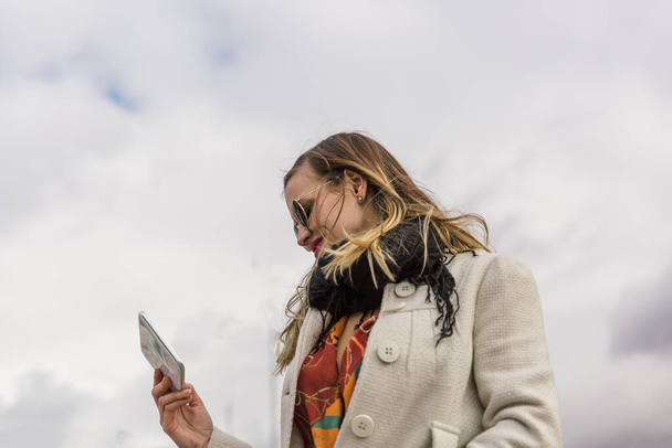 Caucasian blonde girl with white coat and colorful clothes looks at her tablet in the street - Fotoğraf, Görsel