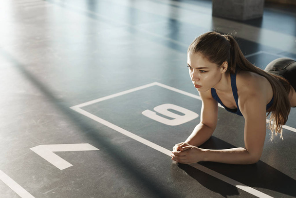 Endurance, sport and healthy lifestyle concept. Side-shot focused young athletic woman, standing in plank on gym floor, looking forward, workout, training to stay fit, morning fitness exercises - Foto, Bild