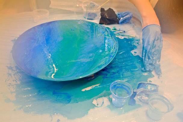 A man in a blue glove beautifully paints draws an acrylic blue multi-colored resin a round home-made board - 写真・画像