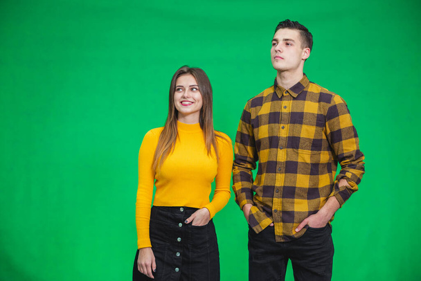 Picture of relaxed couple standing with hands in pockets, posing, weared in trendy clothes on green background. - Zdjęcie, obraz