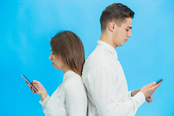 Beautiful young business woman and handsome businessman in formal clothes are using gadgets, standing back to back over blue background. - Φωτογραφία, εικόνα