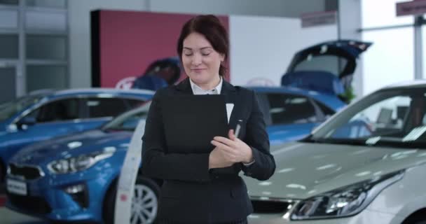 Portrait of adult brunette Caucasian woman with brown eyes posing in car dealership. Smiling dealer in suit holding documents and looking at camera. Cinema 4k ProRes HQ. - Séquence, vidéo