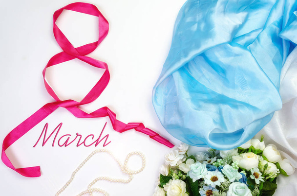 International Women's Day, March 8th. Number eight from a red satin ribbon on an airy satin fabric background with a bouquet of flowers and pink pearls, top view with the inscription March 8 - Фото, изображение