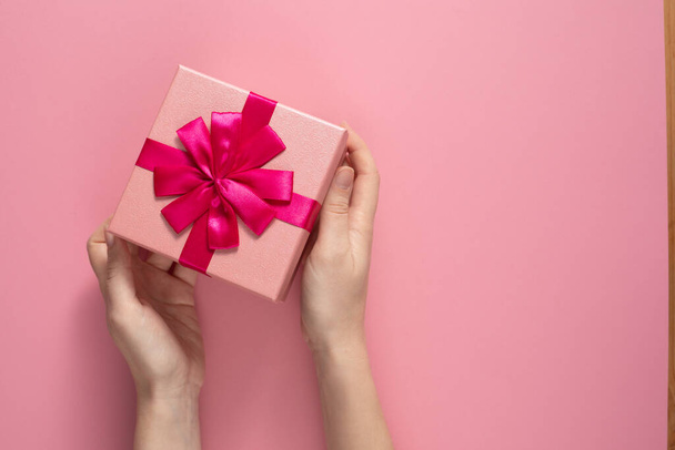Valentine's Day celebration concept. A nice gift from a loved one. Box with a bow in female hands on a delicate pink background. Copy space. Flat lay. - Foto, Imagem