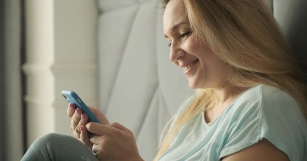 Adult blonde woman using mobile phone, at home. Adult beautiful  woman typing message  on a smartphone. Happy smiling woman uses cell phone lying on bed. Joyful woman holds  smart phone in hands.    - Footage, Video