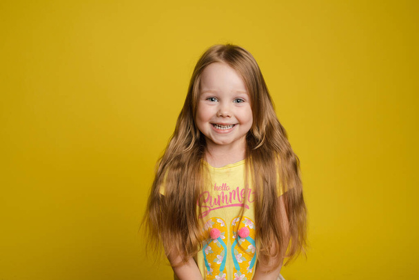 Портрет красивой маленькой девочки с длинными волосами, улыбающейся позируя изолированно на желтом фоне студии
 - Фото, изображение