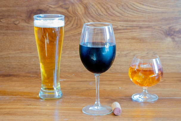 Zusammensetzung der alkoholischen Getränke mit einem Glas Whisky, einem Glas klarem Bier und einem Glas Rotwein auf der Bühne der Eichentische mit Flaschenkorken - Foto, Bild