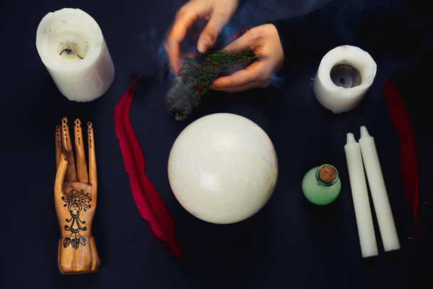 top view flat lay esoteric still life with candles,feather, potion,crystal ball, wooden hand and blurred female hands with smoking juniper - Foto, immagini