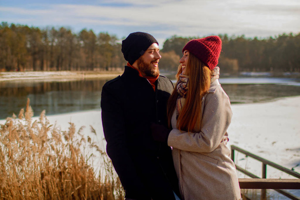 Couple in a winter forest and frozen lake. Young lovers in winter jackets laugh. - Photo, Image
