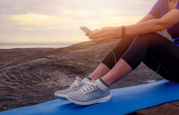 Woman sit and relax on yoga mat and listen to music via smartphone at stone beach by the sea . Woman outdoor workout. Fit girl wear smart band. Healthy, chill out lifestyle. Relax on summer vacations. - Фото, изображение