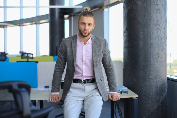 Thoughtful young business man in casual clothes looking at camera and keeping hand on chin while leaning on the desk. - Photo, image