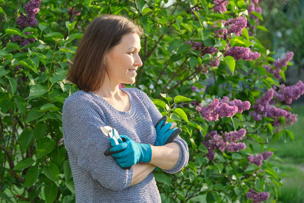 Portrait of mature smiling female gardener in garden gloves with secateurs - 写真・画像