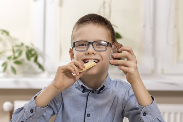 Улыбающийся мальчик в очках сидит за столом и держит сладости в руках. В одной руке он держит белый шоколад, который кусает, а в другой держит шоколадку.
. - Фото, изображение