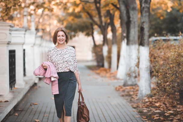 Mujer sonriente de 50-55 años caminando en la calle usando traje sosteniendo el bolso al aire libre. Mirando la cámara. 50 años
.  - Foto, Imagen