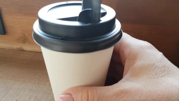 De hand van een vrouw neemt een papieren kopje koffie of thee in een café op een houten tafel met uitzicht op de straat en willekeurige voorbijgangers op een wazige achtergrond. Hand draait het glas - Video