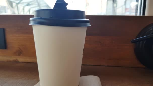 Er staat een witte papieren kop koffie op een houten tafel in het café, en door het glas zie je de straat met een wazige focus. - Video