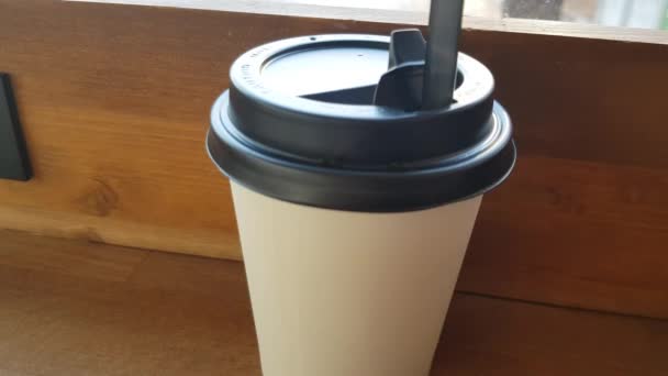 There is a white paper Cup of coffee on a wooden table in the cafe, and through the glass you can see the street with a blurred focus. - Footage, Video