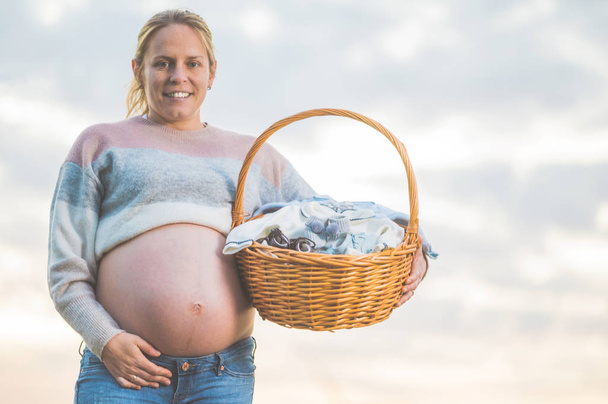 Беременная женщина ждет ребенка, позирует на улице с плетеной корзиной с одеждой для новорожденного
. - Фото, изображение