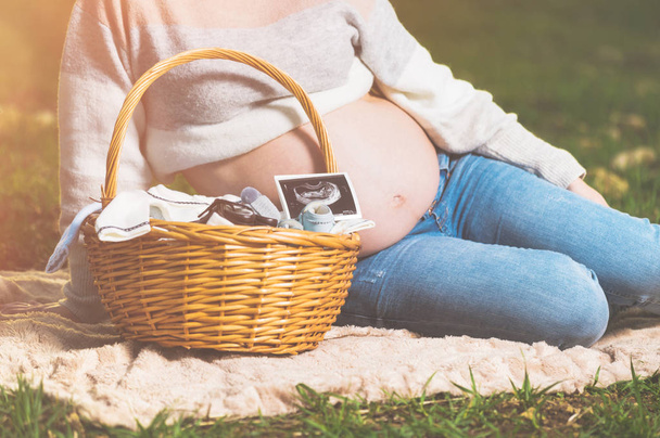 Έγκυος γυναίκα περιμένει μωρό, ποζάρει έξω με ψάθινο καλάθι με ρούχα για το νεογέννητο. - Φωτογραφία, εικόνα