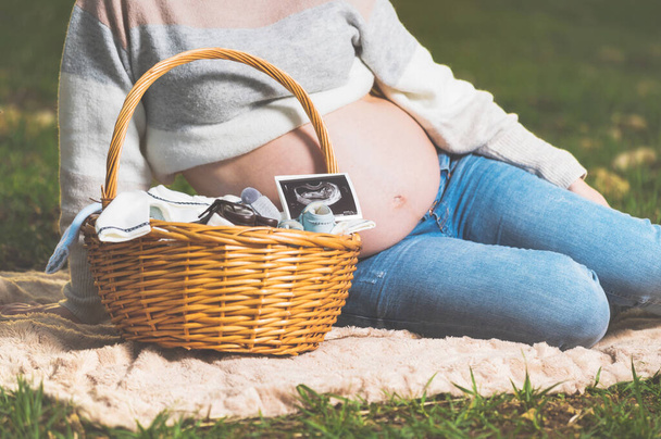 Mujer embarazada esperando un bebé, posando al aire libre con una canasta de mimbre con ropa para su recién nacido
. - Foto, imagen