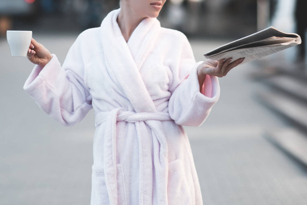 Mujer con albornoz blanco sosteniendo revista fresca y taza de café de pie en la calle al aire libre. Buenos días. Hotel spa resort
.  - Foto, Imagen
