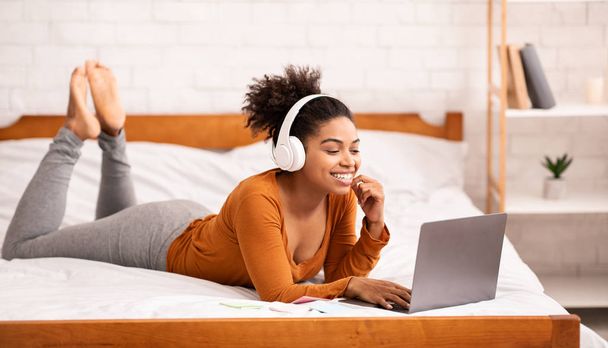 Afro-americana chica en auriculares usando portátil acostado en la cama
 - Foto, imagen
