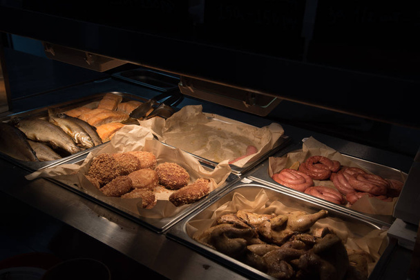 Self-service canteen. Sausages, fish, chicken, and cutlets are laid out in large metal containers - Foto, imagen