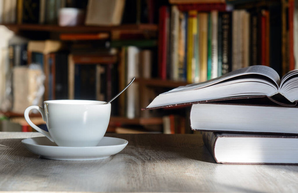 Een witte beker met koffie staat op een tafel verlicht door de zon. Tegenover planken met boeken. Vlakbij is een open boek. Concept - schrijven, koffiepauze, winteravond - Foto, afbeelding