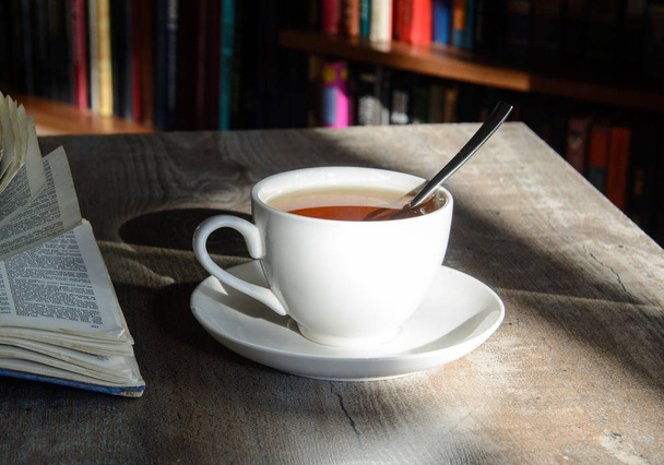 Uma xícara branca com café fica em uma mesa iluminada pelo sol. Prateleiras opostas com livros. Perto está um livro aberto. Conceito - escrita, coffee break, noite de inverno
 - Foto, Imagem