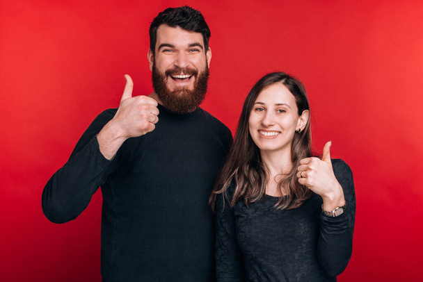 Fotografie radostný mladý pár stojící nad červeným pozadím a ukazující palce nahoru - Fotografie, Obrázek