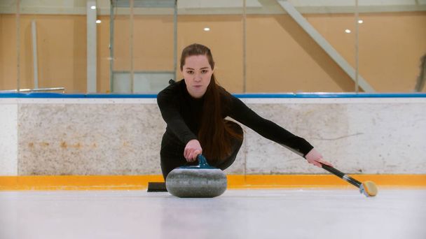 Curling - μια νεαρή γυναίκα με μακριά μαλλιά στο παγοδρόμιο κρατώντας μια πέτρα γρανίτη και κρατώντας ένα πινέλο - Φωτογραφία, εικόνα