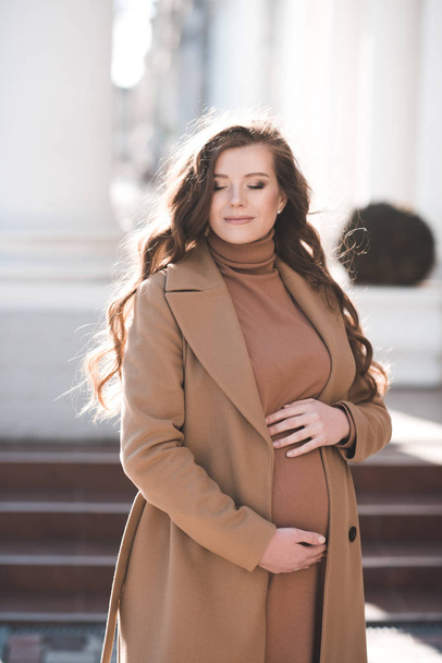 Hymyilevä onnellinen raskaana oleva nainen 25-26 vuotias tilalla kädet hänen vatsa yllään tyylikäs beige takki ulkona kaupungin kadulla. Äitiys. Parikymppinen Kevätkausi
.  - Valokuva, kuva