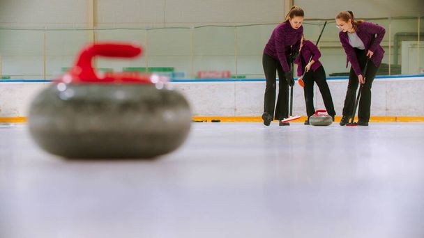 Curling training - leidende granieten steen op het ijs - twee vrouwen wrijven het ijs voor de steen - Foto, afbeelding