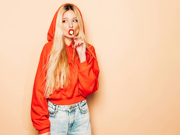 Portrait of young beautiful hipster bad girl in trendy red summer hoodie and earring in her nose.Sexy carefree smiling blond woman posing in studio.Positive model licking round sugar candy - Foto, afbeelding