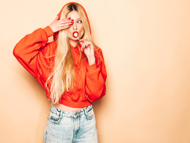 Portrait of young beautiful hipster bad girl in trendy red summer hoodie and earring in her nose.Sexy carefree smiling blond woman posing in studio.Positive model licking round sugar candy - 写真・画像