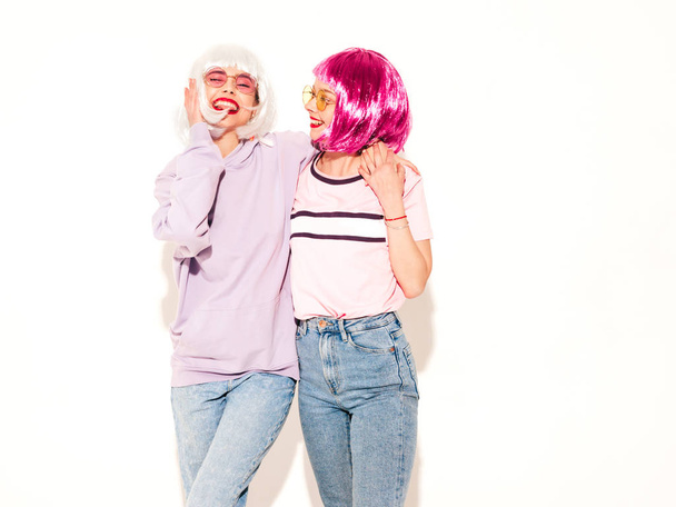 Two young sexy smiling hipster girls in wigs and red lips.Beautiful trendy women in summer clothes.Carefree models posing near white wall in studio.Positive female going crazy and hugging - Photo, Image
