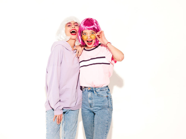 Two young sexy smiling hipster girls in wigs and red lips.Beautiful trendy women in summer clothes.Carefree models posing near white wall in studio.Positive female going crazy and hugging - Фото, зображення