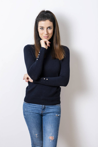 Portrait shot of beautiful woman with hand on chin wearing turtleneck sweater and blue jeans while standing at isolated white background.  - Foto, Imagen