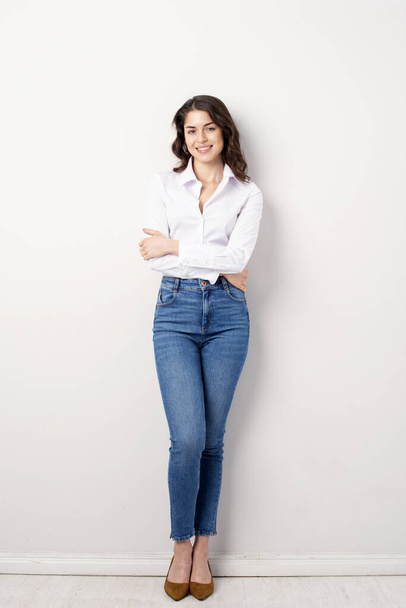 Full length studio shot of happy young woman wearing white shirt and blue jeans while posing at isolated white background. - Фото, изображение