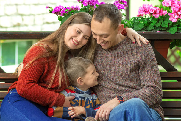 Ευτυχισμένη οικογένεια που κρατά τα χέρια κάθεται και ποζάρει σε ένα παγκάκι στο υπαίθριο πάρκο, έχοντας ποιοτικό χρόνο μαζί. - Φωτογραφία, εικόνα