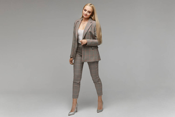 Smiling young elegant businesswoman with bright makeup and in suit looking down in the studio, isolated on grey background. Model girl in the business outfit, portrait in full length with copy space. - Zdjęcie, obraz