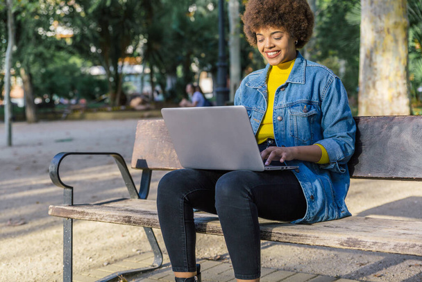Young girl with afro hair in blue denim shirt sitting on a bench working with her laptop in a park - Fotó, kép