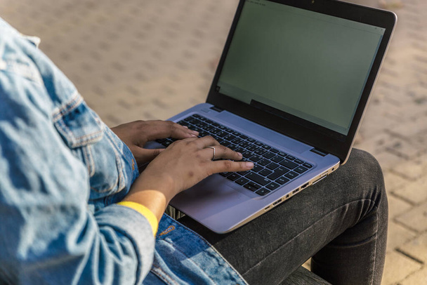 View of the hands of a woman typing on her laptop sitting on a park bench - Foto, afbeelding